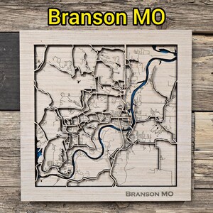 May include: A wooden map of Branson, Missouri, with a detailed laser-cut design. The map features the city's layout, with a blue river and the text "Branson MO" at the top and bottom. The map is set in a light-colored wooden frame.