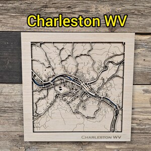 May include: A wooden map of Charleston, WV, with intricate laser-cut details. The map features the city's layout, including the river and streets, with the text "Charleston WV" at the top and bottom. The map is set in a light-colored wooden frame.