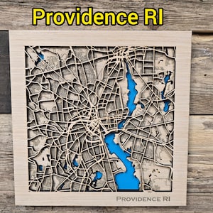 May include: A wooden map of Providence, RI, with a light wood frame. The city's streets and waterways are intricately cut out, with blue representing the water. The text "Providence RI" is at the top and bottom of the frame.