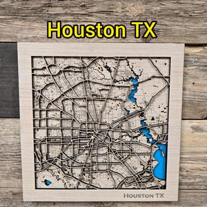 May include: A wooden map of Houston, TX, with intricate laser-cut details. The city's layout is depicted in dark brown, with blue accents representing waterways. The text "Houston TX" is displayed at the top and bottom of the map.