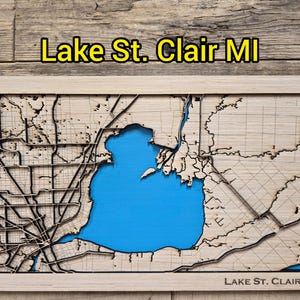 May include: A wooden map of Lake St. Clair, MI, with a blue lake and surrounding land. The map is laser-cut with intricate details of roads and waterways. The text "Lake St. Clair MI" is displayed at the top and bottom of the map.