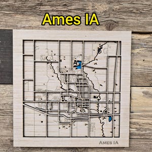 May include: A wooden map of Ames, Iowa, with a light wood frame. The city's layout is intricately carved, with blue accents for water features. The text "Ames IA" is displayed at the top and bottom of the map.
