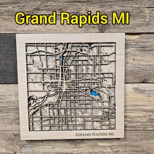 Może przedstawiać: Drewniana mapa Grand Rapids, MI, wycięta laserowo, ze szczegółowymi ulicami i wodą. Nazwa miasta jest wyświetlana na żółto nad i pod mapą. Mapa jest na tle rustykalnego drewna.