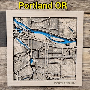 May include: A wooden map of Portland, Oregon, with a light wood frame. The city's layout is intricately carved, with the Willamette River highlighted in blue. The text "Portland OR" is displayed at the top and bottom of the frame.