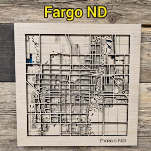 May include: A wooden map of Fargo, ND, with intricate laser-cut details. The map features the city's layout, including streets, buildings, and the Red River. The text "Fargo ND" is at the top and bottom of the map.