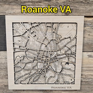 May include: A wooden map of Roanoke, VA, with a detailed street layout. The map is laser-cut and framed in a light wood. The text "Roanoke VA" is displayed at the top and bottom of the map.