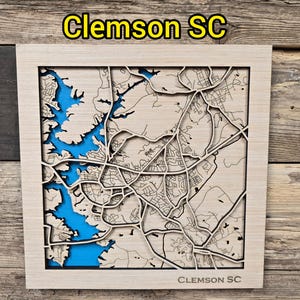 May include: A wooden map of Clemson, SC, with a detailed laser-cut design. The map features a blue-colored lake and river system, with the city's streets and landmarks intricately carved. The text "Clemson SC" is displayed on the top and bottom of the frame.