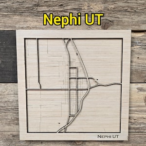 Puede incluir: Un mapa de madera de Nephi, Utah, con el diseño de la ciudad tallado en la madera. El texto "Nephi UT" se muestra en la parte superior e inferior del mapa. El mapa está sobre un fondo de madera rústica.