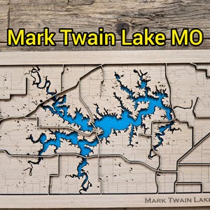 May include: A laser-cut wooden map of Mark Twain Lake, MO, featuring a blue lake and surrounding land. The map is framed in a light wood colour, with the lake's water a vibrant blue. The text "Mark Twain Lake MO" is displayed at the top and bottom.