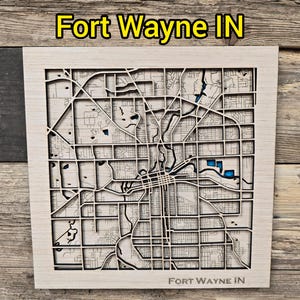 May include: A wooden map of Fort Wayne, Indiana, with intricate laser-cut streets and waterways. The map features a light wood finish with blue accents for the water areas. The text "Fort Wayne IN" is displayed at the top and bottom.