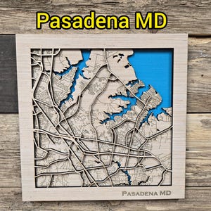 Könnte beinhalten: Eine Holzkarte von Pasadena, MD, in einem hellen Holzrahmen. Die Karte zeigt detaillierte, lasergeschnittene Straßen und Wasserwege, mit einem blauen Hintergrund für das Wasser. Der Text "Pasadena MD" ist sichtbar.