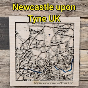 May include: A laser-cut wooden map of Newcastle upon Tyne, UK. The map shows the city's layout, with streets and the River Tyne in blue. The text "Newcastle upon Tyne UK" is displayed at the top and bottom. The map is set in a wooden frame.