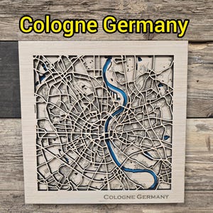 Puede incluir: Un mapa de madera de Colonia, Alemania, con un diseño detallado de las calles y un río azul. El nombre de la ciudad se muestra en la parte superior e inferior del marco. El mapa está sobre un fondo de madera rústica.