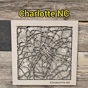 May include: A wooden map of Charlotte, NC, with intricate laser-cut streets and buildings. The city name is displayed in yellow text at the top and bottom of the frame. The map is set against a rustic wooden background.