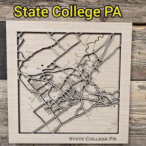 May include: A wooden map of State College, PA, with intricate laser-cut details. The map features roads, buildings, and natural features, all within a square frame. The text "State College PA" is visible at the top and bottom of the frame.