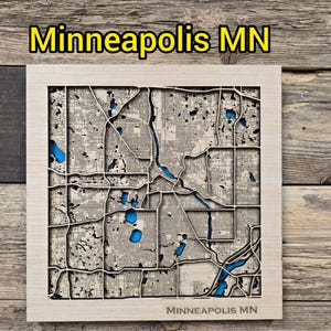 May include: A wooden map of Minneapolis, MN, with a detailed laser-cut design. The map features the city's layout, including streets, parks, and the Mississippi River, with blue accents for lakes and waterways. The text "Minneapolis MN" is displayed at the top and bottom.
