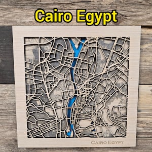 May include: A wooden map of Cairo, Egypt, with a detailed laser-cut design. The map features a blue river and city streets, set within a light-colored wooden frame. The text "Cairo Egypt" is displayed at the top and bottom of the frame.