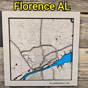 May include: A wooden map of Florence, AL, with a light-colored frame. The map features a detailed street layout and a blue river. The text "Florence AL" is displayed at the top and bottom of the frame.