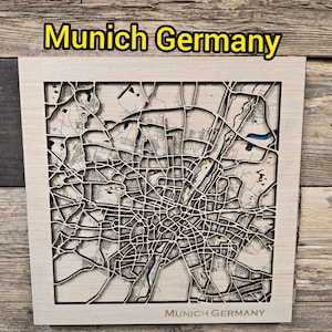 Puede incluir: Un cuadro de madera cuadrado con un mapa multicapa cortado con láser de Múnich, Alemania. El marco de madera clara rodea intrincados patrones de calles en madera oscura, con pequeños detalles en azul que representan cuerpos de agua.