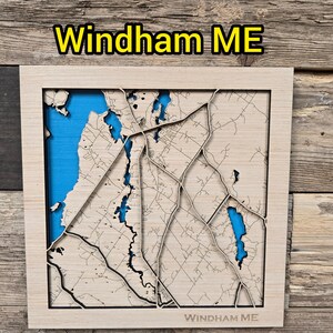 May include: A wooden map of Windham, ME, with a light wood frame. The map features a detailed laser-cut design with blue water features. The text "Windham ME" is displayed at the top and bottom of the frame.