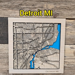May include: A wooden map of Detroit, MI, with a light wood frame. The city's layout is intricately carved, with blue representing the water areas. The text "Detroit MI" is displayed at the top and bottom of the map.