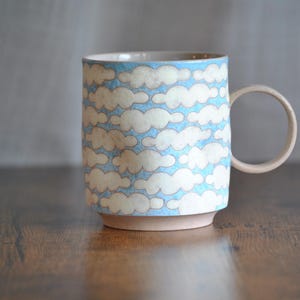 May include: A ceramic mug featuring a repeating pattern of off-white clouds against a light blue background. The mug has a cylindrical body, a circular handle on the right side, and a plain, unglazed base that shows the natural clay color.
