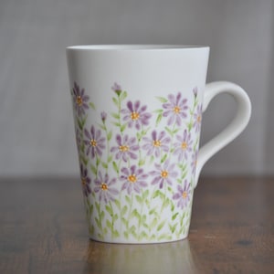 May include: White ceramic mug decorated with purple and yellow floral design. The flowers are painted with a watercolor effect, and the mug has a curved handle. The mug is a kitchen and dining item.
