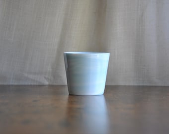 May include: A light blue ceramic cup with a slightly flared shape. The cup is set on a dark brown wooden surface, with a neutral fabric backdrop. The cup's design features subtle horizontal stripes.