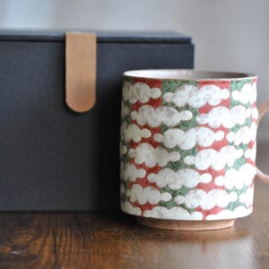 Puede incluir: Una taza de cerámica con un diseño de nubes en blanco, rojo y verde. La taza tiene un asa y una base de color marrón claro. Detrás de la taza hay una caja negra con una correa de cuero. La taza está sobre una superficie de madera.