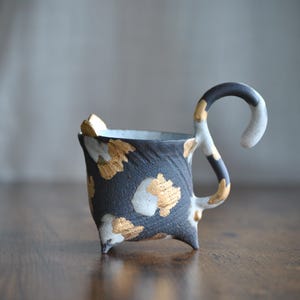 May include: A handcrafted ceramic mug with a unique design. The mug features a dark gray base adorned with gold and white accents. The handle is a curved shape with a black, white, and gold color scheme. The mug sits on three small feet.