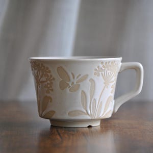 Könnte beinhalten: Cremefarbene Keramiktasse mit geprägten Blumen- und Schmetterlingsmotiven. Die Tasse hat einen quadratischen Griff und einen leicht ausgestellten Boden. Das Design zeigt detaillierte botanische und Insektenmotive und verleiht dem Geschirr einen Hauch von naturnaher Eleganz.