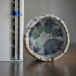 以下が含まれることがあります： ユニークな有機的な形状の、手作りの小さなセラミックボウル。内側には緑と青の花柄のデザインが施されています。縁は薄茶色です。ボウルの直径は約7.6cmです。