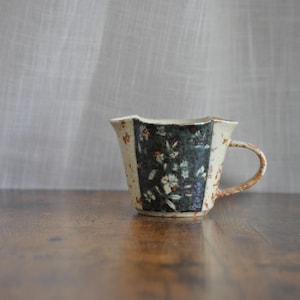 May include: A ceramic mug with a unique, irregular shape. The mug features a dark teal panel with white floral designs, contrasted by cream-colored sections. The handle and edges have a speckled, rustic appearance, suggesting a handmade quality.