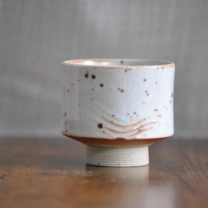 Può includere: Una tazza in ceramica bianca con macchie marroni e una superficie testurizzata. La tazza ha una base larga e una parte superiore arrotondata.