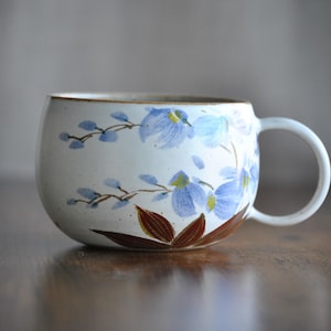 Könnte beinhalten: Eine weiße Keramiktasse mit runder Form und Henkel. Die Tasse ist mit blauen Blumenmustern und braunen Blättern verziert. Der Rand hat eine goldfarbene Verzierung. Die Tasse steht auf einer Holzoberfläche.