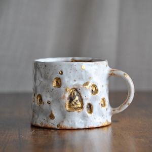 May include: A handmade ceramic mug with a textured, off-white surface. The mug features gold-colored accents, including small, recessed designs. The handle is rounded, and the mug sits on a wooden surface. A unique piece for coffee or tea.