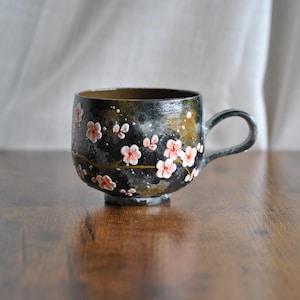 May include: A black ceramic mug with a handle, adorned with delicate pink and white floral designs. The mug sits on a wooden surface, showcasing its unique, handcrafted appearance. The cup is approximately 4 inches in height.