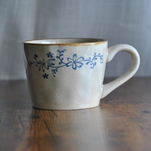 May include: A beige ceramic mug with a speckled finish and a blue floral vine pattern encircling the upper portion. The mug features a sturdy, curved handle on the right side and a slightly flared rim with a subtle brown edge detail.