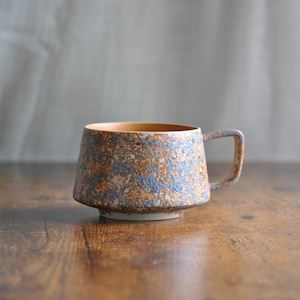 Może przedstawiać: Ceramiczny kubek o unikalnej, teksturowanej powierzchni. Kubek ma nakrapiany wzór w odcieniach niebieskiego i brązowego, z jasnobrązowym wnętrzem. Ma kwadratowy uchwyt i stoi na małej, okrągłej podstawie. Kubek ma około 10 cm wysokości.