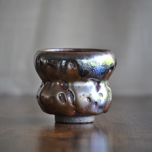 May include: A handcrafted ceramic sake cup with a unique, textured surface and a dark, glossy glaze. The cup has a bulbous shape with a small, circular base. The colors are primarily brown and black, with hints of blue and green.