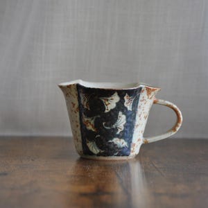 May include: A ceramic mug with a unique, irregular shape and a handle. The mug features a cream-colored base with a dark blue panel adorned with white floral patterns. The rim and handle have a rustic, brown finish. The mug is on a wooden surface.