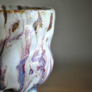 以下が含まれることがあります： ユニークな大理石模様の手作りセラミックカップ。カップは、茶色、赤、青の縞模様の白いベースが特徴です。カップの形はわずかに不規則で、手作りの魅力を加えています。このカップは飲み物に適しています。