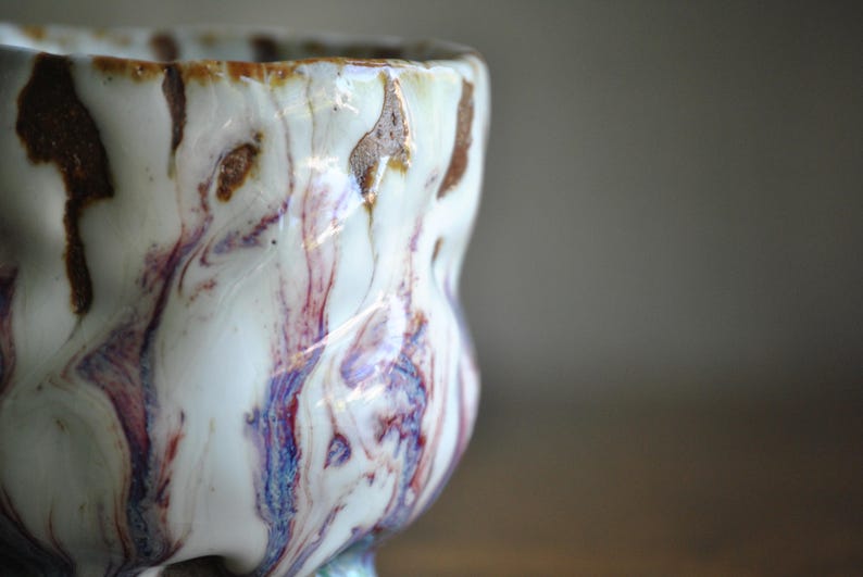 以下が含まれることがあります： ユニークな大理石模様の手作りセラミックカップ。カップは、茶色、赤、青の縞模様の白いベースが特徴です。カップの形はわずかに不規則で、手作りの魅力を加えています。このカップは飲み物に適しています。
