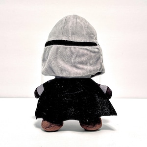 May include: A plush toy depicting a character from a popular science fiction franchise. The toy has a gray helmet with a black band, a black cape, and brown feet. The toy is designed to resemble a character from the Star Wars franchise.