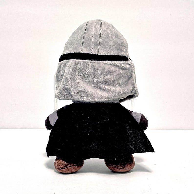 May include: A plush toy depicting a character from a popular science fiction franchise. The toy has a gray helmet with a black band, a black cape, and brown feet. The toy is designed to resemble a character from the Star Wars franchise.