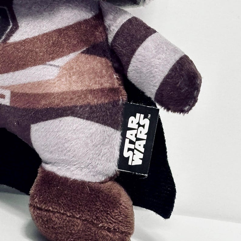 May include: A plush toy featuring a brown and gray color scheme with a black cape. The Star Wars logo is visible on a black tag attached to the toy. The toy has a soft, textured appearance.