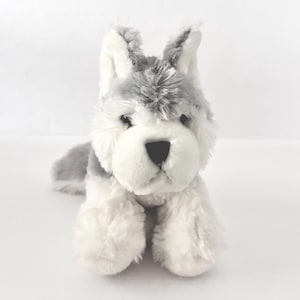 May include: A plush toy dog in a lying position, featuring a soft, fluffy texture. The toy has a white and gray color scheme, with a white muzzle and paws, and gray fur on the head and back. The dog has black eyes and a black nose.