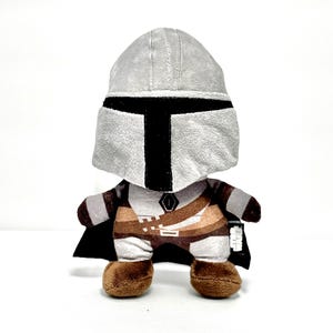 May include: A plush toy depicting a character from a popular science fiction series. The toy features a gray helmet with a black visor, a brown and tan outfit, and a black cape. The toy is standing on a white surface.