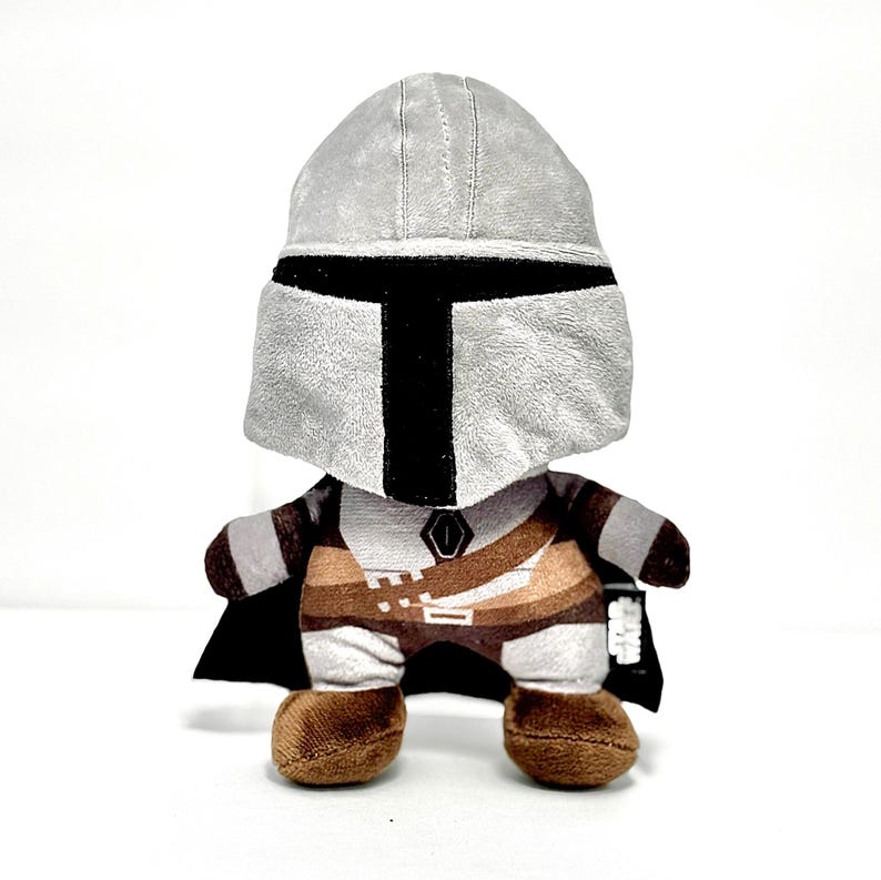May include: A plush toy depicting a character from a popular science fiction series. The toy features a gray helmet with a black visor, a brown and tan outfit, and a black cape. The toy is standing on a white surface.