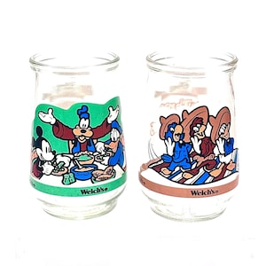 May include: Two clear glass Welch's jelly jars with colorful Disney character graphics. One jar features Mickey Mouse, Goofy, and Donald Duck at a table. The other jar shows three characters in sombreros. Both jars have a green and brown band at the base.